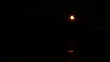 Full-moon-reflection-in-small-waves-on-water-surrounded-by-deep-black-background,-small-city-lights-in-the-distance
