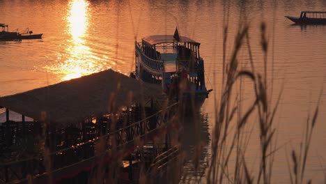 Medium-Exterior-Shot-of-A-Boat-Coming-in-Around-Another-Boat-on-a-Lake-With-the-Reflection-of-the-Sun-in-Golden-Hour-Through-Some-Grass