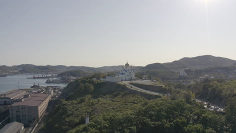 Slow-pivot-aerial-shot-of-an-orthodox-church-with-blue-roof-and-golden-domes,-located-on-top-of-the-hill-with-port-and-city-building-in-the-background,-on-a-bright,-clear,-sunny-day