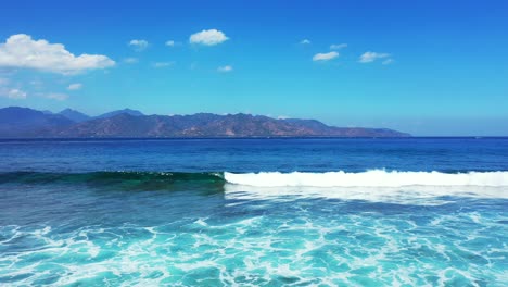 Beautiful-white-waves-splashing-on-shore-of-tropical-islands-surrounded-by-blue-sea-on-a-bright-sky-with-clouds-background,-Indonesia