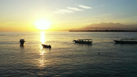 Tranquil-bay-with-anchored-boats-near-shore-of-tropical-island-at-sunset-yellow-sky-reflecting-over-mountains-horizon,-Bali