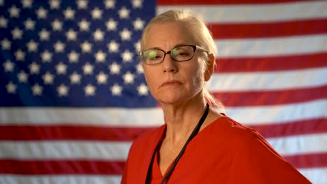 Medium-tight-portrait-of-the-back-of-blonde-nurses-head-as-she-turns-and-looks-at-camera-looking-angry-wearing-glasses-with-out-of-focus-US-flag