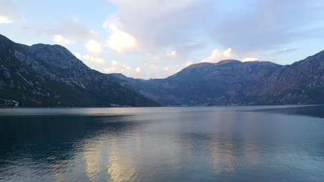 The-bay-of-Kotor-in-Montenegro,-with-its-still-waters,-surrounded-by-high-mountain-range,-reflected-in-the-water,-white-clouds-above-the-horizon,-late-afternoon-sunlight-shining-over-the-peaks