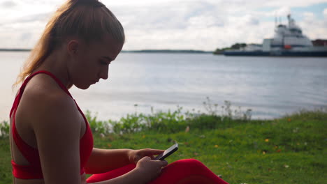 Junges-Mädchen-In-Roter-Sportbekleidung,-Das-Auf-Dem-Gras-Am-Meer-Sitzt-Und-Auf-Dem-Smartphone-Scrollt