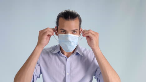 Happy-Adult-Male-with-Hazel-Eyes-Wearing-Collared-Button-Down-Shirt-Puts-on-Surgical-Mask-on-a-Gray-Background-to-Protect-from-Covid-19---Coronavirus,-Straight-on-Close-Up-Tripod-Shot