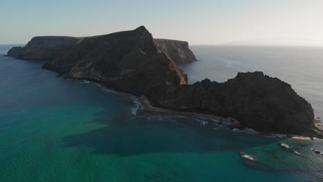 Breathtaking-scenic-summer-flight-above-turquoise-ocean-sea-waves-rolling-on-Calheta-sandy-beach-toward-picturesque-Cal-Islet-dramatic-brown-island-and-mountain-peak-at-sunset,-aerial-approach