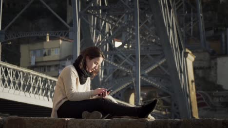 Gorgeous-Lady-Using-Smartphone-While-Sitting-By-The-Steel-Bridge-Structure-In-Porto,-Portugal---full-slowmo-shot