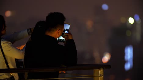 Dos-Mujeres-Asiáticas-Y-Un-Hombre-Están-Tomando-Fotos-Con-Sus-Teléfonos-Celulares-Del-Horizonte-De-La-Ciudad-Por-La-Noche,-Toma-De-Mano-Con-Zoom