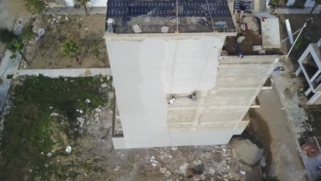 Concrete-plaster-finish-applied-to-building-side-in-Sam-Son-Vietnam-as-two-construction-workers-swing-back-and-forth,-Aerial-top-view-shot