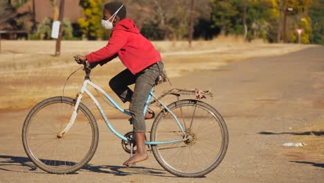 Young-black-teenager-rides-his-bike-down-a-dirt-path,-wearing-face-mask-but-no-shoes,-South-Africa