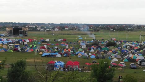 Flying-Over-Trees-Revealing-Multi-Colored-Tents-and-Stage-in-a-Field-at-a-Music-Festival-With-Town-in-Background