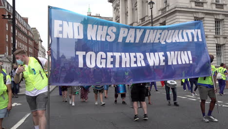 Hundreds-of-National-Health-Service-staff-and-key-workers-march-with-a-large-blue-banner-that-says,-“End-NHS-Pay-Inequality