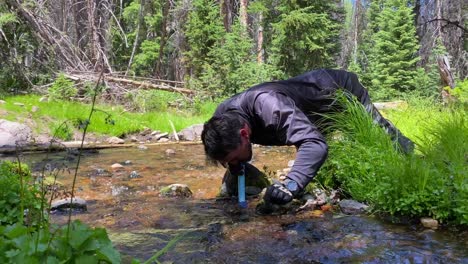 Man-wearing-dirt-bike-gear-drinking-water-from-a-wilderness-stream-using-a-life-straw-clean-water-filter-system