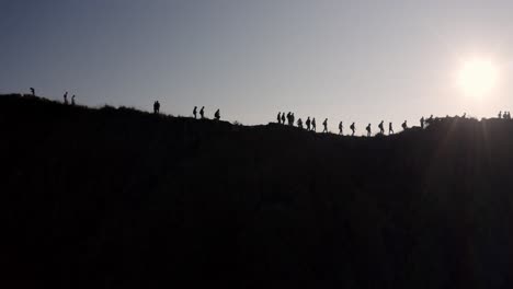 Silhouetted-People-at-the-summit-of-Mout-Batur-volcano-in-Bali-Indonesia-walk-along-the-crater-ridge,-Aerial-pan-right-shot