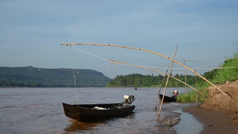 Long-Boats-with-motors-moored-and-bouncing-on-Mekong-River-as-a-boatman-speeds-up-its-longboat-towards-the-left-side-of-the-frame-with-mountain-background-and-lovely-bluesky