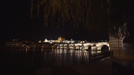 The-Prague-Castle-and-Charles-bridge-over-river-Vltava-in-the-historical-centre-of-Prague,Czechia,lit-by-lights-at-night,viewed-from-the-other-side-of-the-river,pedestal-shot-from-under-a-willow-tree