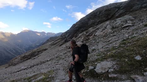 A-young,-fit-man-with-long-hair-and-a-large-backpack-is-jogging-and-running-carefully-down-a-path-in-themountains-full-of-rocks-and-stones