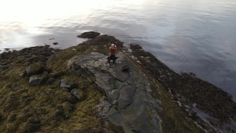 Aerial-footage-circling-around-a-man-standing-at-the-shore-in-Trondheim,-Norway-and-taking-pictures-with-a-camera