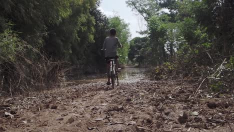 Young-boy-trying-to-Cycle-with-old-bike-through-flooded-rural-road-after-heavy-rain-in-Cambodia