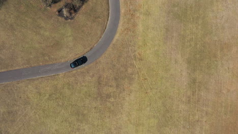 An-aerial,-top-down-drone-view-over-a-single-car-driving-through-an-empty-park-on-a-sunny-day