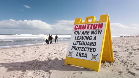 Surfers-Walk-by-Ocean-Behind-Beach-Caution-Sign-on-Sunny-Florida-Day