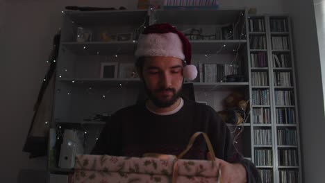 Male-speaking-on-videocall,-opening-a-Christmas-present-while-wearing-a-Christmas-hat