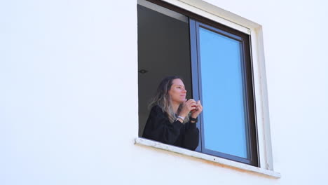 Medium-shot-of-young-blond-woman-on-window-drinking-coffee-or-tea-and-waving-at-neighbor