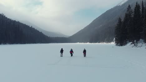 aerial-shot-of-a-group-of-people-walking-on-a-frozen-lake-surrounded-by-pine-trees-and-mountains-during-winter-time,-British-Columbia-Canada