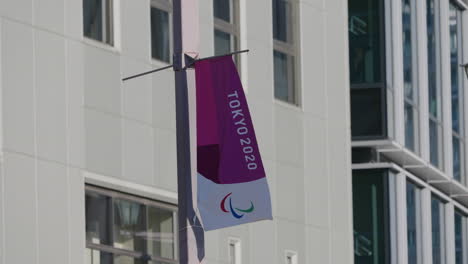 Bandera-Olímpica-De-Tokio-2020-Ondeando-Y-Ondeando-En-El-Viento-Con-Edificios-En-El-Fondo---Tiro-De-ángulo-Bajo