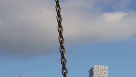 Revealing-close-up-of-yellow-metal-industrial-chain-and-hook-to-lift-cargo-anchor-and-secure-boats-in-the-marina-with-a-blurry-cityscape-in-the-background-blue-sky-clouds-buildings-boats