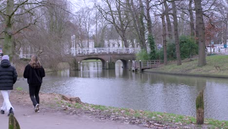 Dos-Amigos-Caminando-Juntos-Por-Un-Parque,-A-Lo-Largo-Del-Canal-En-Utrecht,-Países-Bajos,-Con-Un-Hermoso-Puente-En-El-Fondo