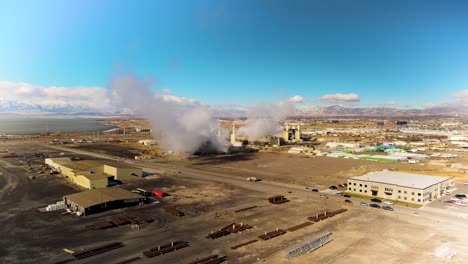An-industrial-complex-emits-smoke-and-steam-into-the-air-with-an-aerial-view-of-a-lake-and-snowy-mountains