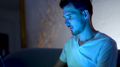 A-young-man-with-a-stubble-and-a-modern-hairstyle,-wearing-a-white-t-shirt,-sitting-at-home-on-a-sofa,-working-on-his-laptop-late-at-night,-lit-by-its-blue-light,-static-close-up-4k