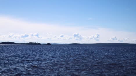 Zeitraffer-Des-Fließenden-Wassers-Eines-Riesigen-Sees-In-Finnland-Mit-Weißen-Wolken,-Die-In-Der-Ferne-über-Etwas-Vegetation-Vorbeiziehen