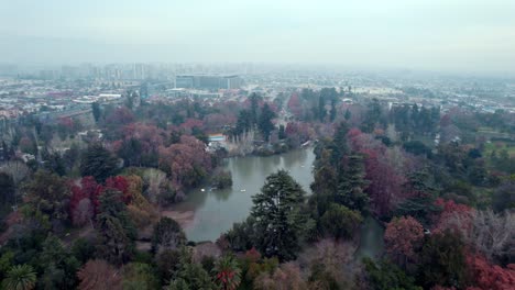 Aerial-dolly-in-of-artificial-pond-surrounded-by-colorful-autumnal-trees-and-vegetation-in-O'Higgins-Park,-smoggy-day,-Santiago,-Chile