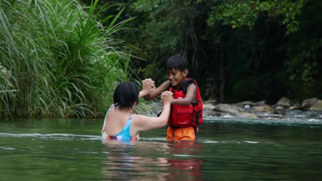 Indian-boy-wearing-red-life-jacket-playfully-hand-wrestling-with-Asian-woman-in-river
