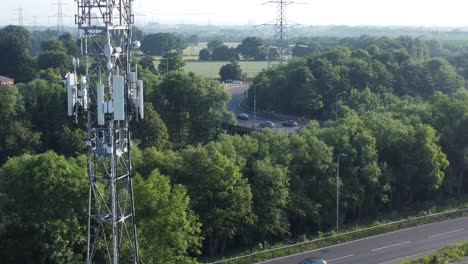 5G-broadcasting-tower-antenna-in-British-countryside-with-vehicles-travelling-on-highway-background-aerial-rising-slow-view