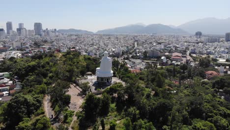 Tourist-attraction-of-white-Buddha-statue-in-park-and-cityscape-of-Nha-Trang,-beautiful-sunny-day-in-Vietnam---aerial-panoramic