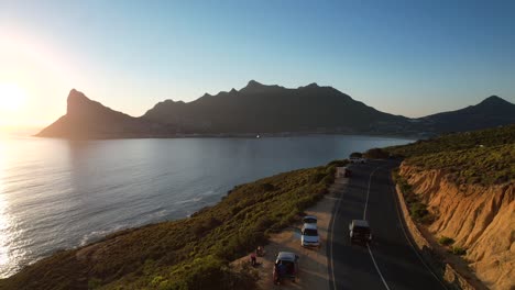 Auto-Fährt-Entlang-Der-Küste-Von-Hout-Bay-Mit-Sonnenuntergang-über-Dem-Sentinel-Peak-In-Kapstadt,-Luftaufnahmen