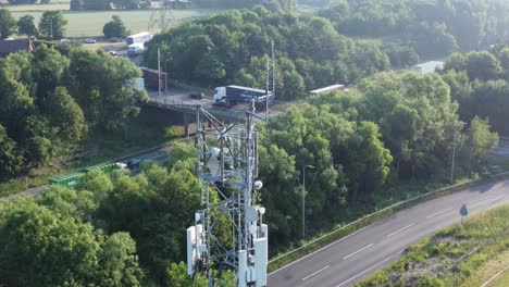 5G-broadcasting-tower-antenna-in-British-countryside-with-vehicles-travelling-on-highway-background-aerial-tilt-up-view