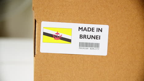 Hands-applying-MADE-IN-BRUNEI-flag-label-on-a-shipping-cardboard-box-with-products