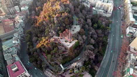 Aerial-view-of-Caupolican-Terrace-at-Santa-Lucia-hill-covered-in-autumnal-trees,-traffic-in-avenues-at-sunset,-Santiago,-Chile