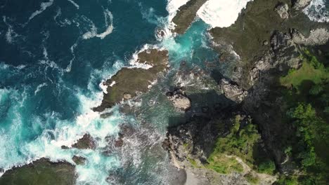 Sliding-overhead-shot-of-waves-of-Indian-Ocean-hitting-boulder-and-coral-reef-in-the-beach-in-sunny-day,-seen-hill-overgrown-with-trees-on-the-side---Pengilon-Hill,-Indonesia,-Asia