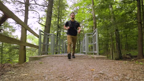 Male-hiker-with-backpack-walks-quickly-up-metal-staircase-towards-camera-in-forest-landscape,-low-angle-view