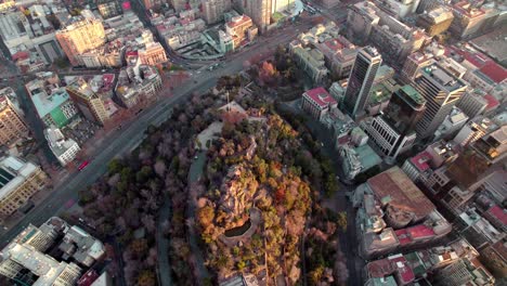 Aerial-dolly-in-of-Santa-Lucia-Hill-covered-in-autumnal-trees,-traffic-in-Alameda-avenue-and-Santiago-buildings-in-back,-Chile