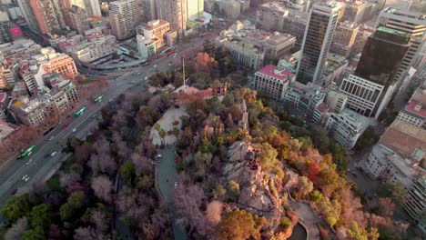 Aerial-orbit-of-Santa-Lucia-Hill-with-autumnal-trees,-traffic-in-Alameda-avenue,-Santiago-city-buildings-in-back-at-sunset,-Chile