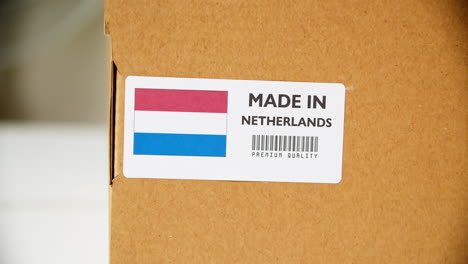 Manos-Aplicando-Etiqueta-De-Bandera-Hecha-En-Holanda-En-Una-Caja-De-Cartón-De-Envío-Con-Productos