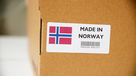 Hands-applying-MADE-IN-NORWAY-flag-label-on-a-shipping-cardboard-box-with-products