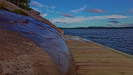 Shot-of-a-wooden-pier-along-rocky-slope-beside-a-calm-lake-throughout-the-day-in-timelapse