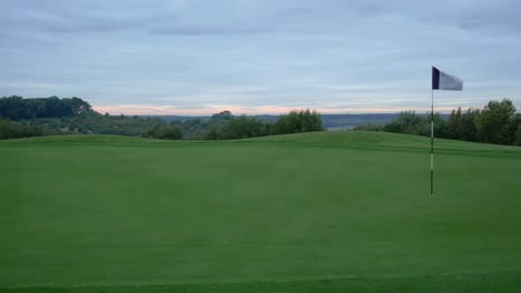 Schwenkende-4K-Aufnahme-Eines-Golfplatzgrüns-Und-Einer-Flagge-Bei-Sonnenaufgang